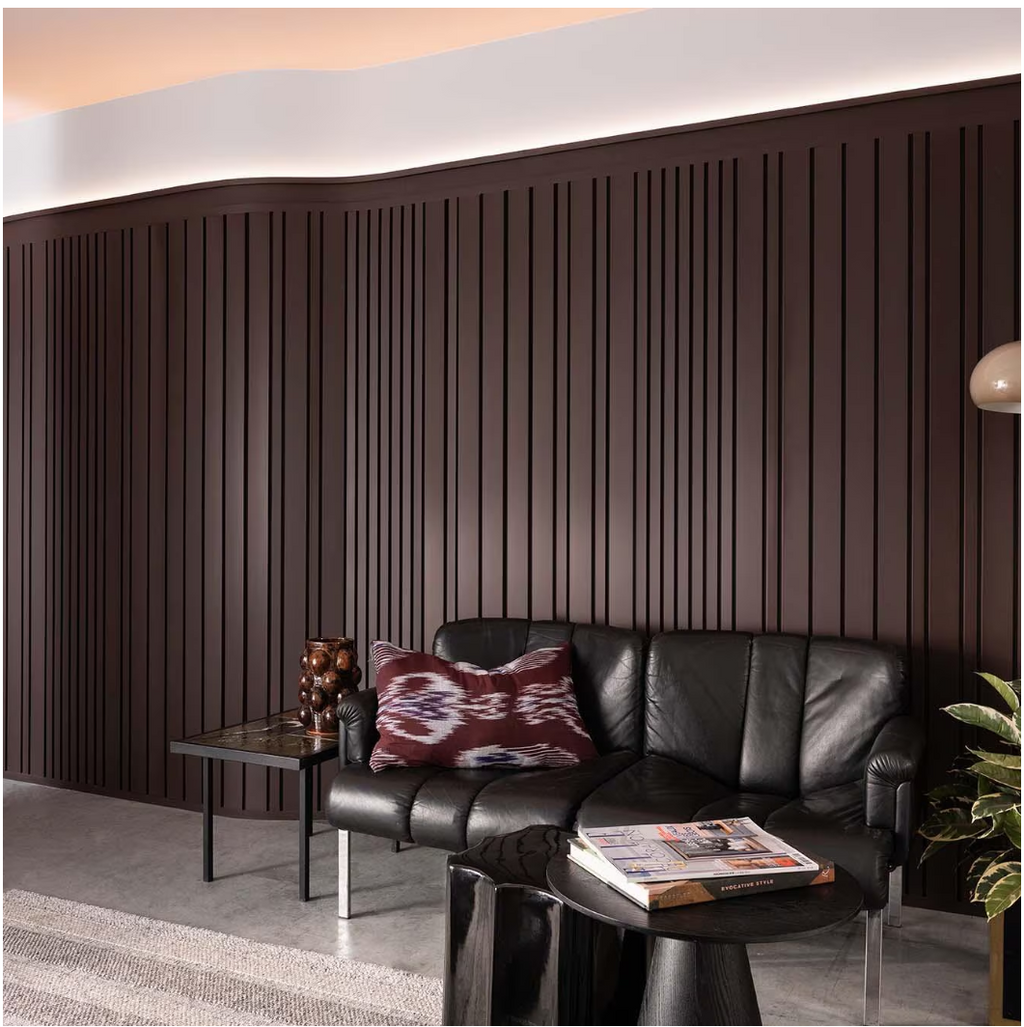 Floor-to-ceiling curved wall panels with vertical grooves in deep mahogany behind a modern black leather couch in a softly lit living room.