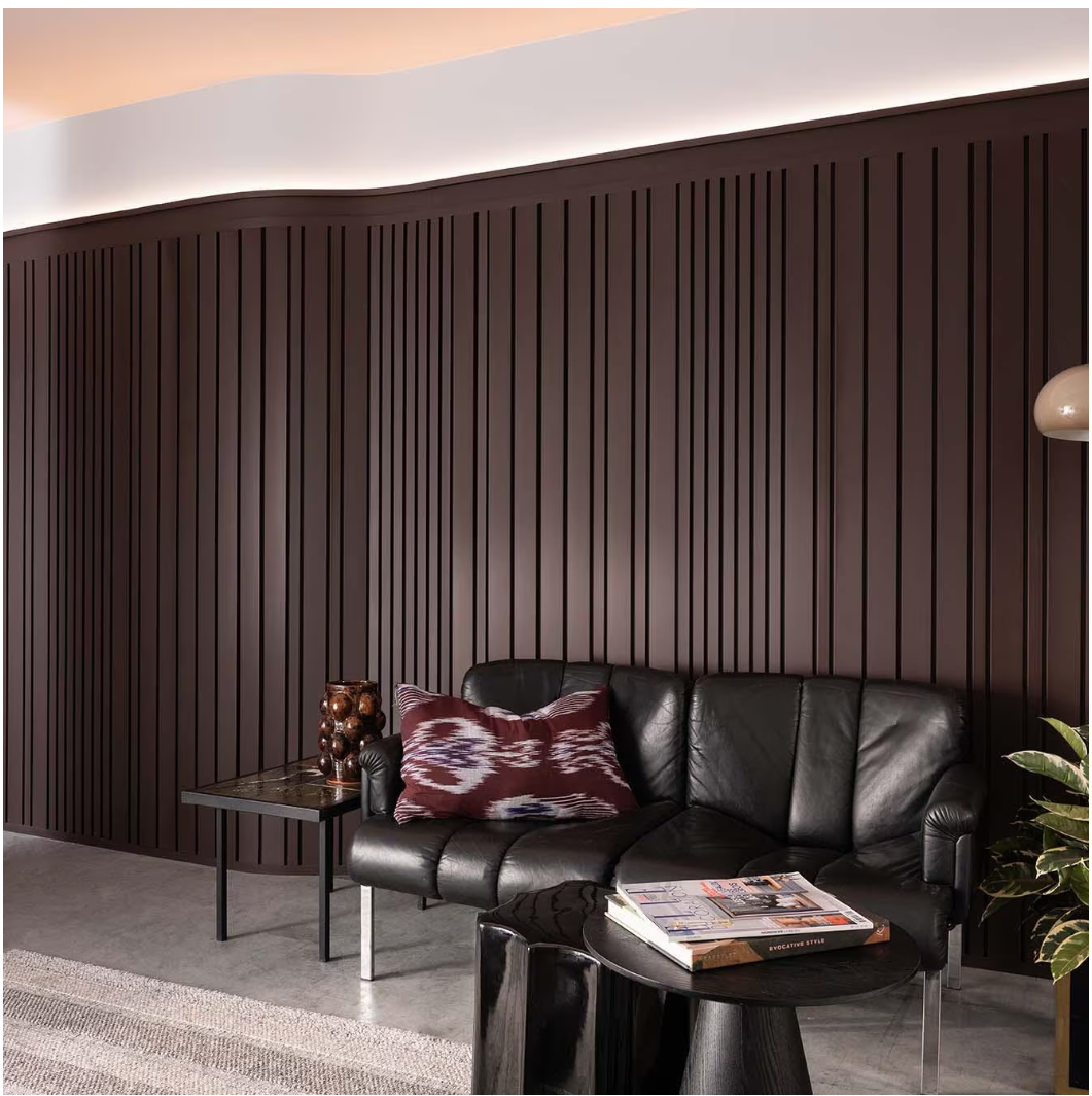 Floor-to-ceiling curved wall panels with vertical grooves in deep mahogany behind a modern black leather couch in a softly lit living room.